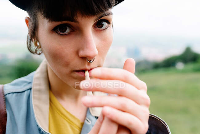 Crop informal lady in casual clothes with piercing in nose lighting cigarette while standing on street and looking at camera — Stock Photo
