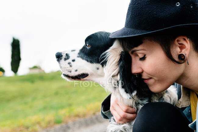 Joven mujer alegre con ropa casual y sombrero sentado en el suelo abrazando a su perro durante el paseo por la calle. - foto de stock