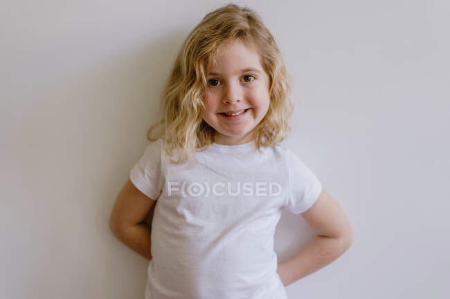 Позитивный ребенок в повседневной одежде улыбается и смотрит в камеру, стоя и опираясь на белую стену в современной студии — стоковое фото