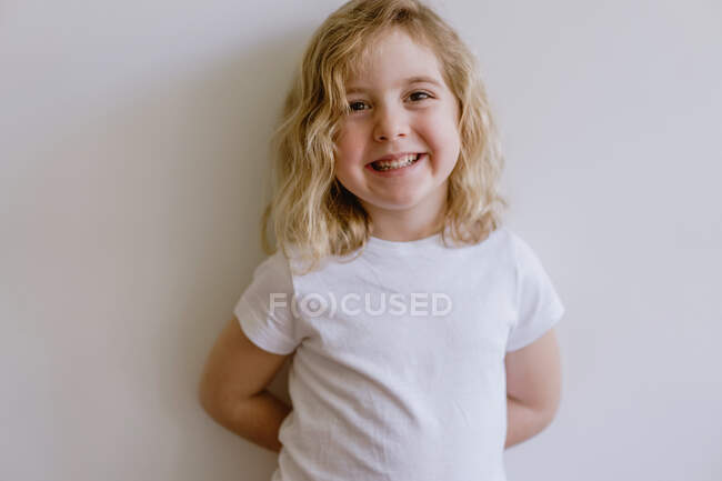 Positive child wearing casual clothes smiling and looking at camera while standing and leaning on white wall in modern studio — Stock Photo