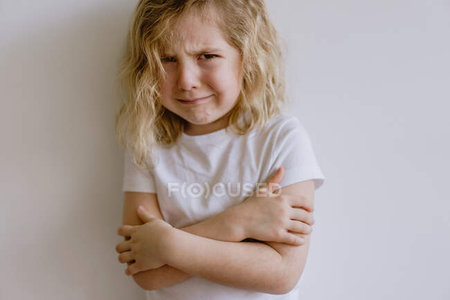 Непослушный ребенок с волнистыми волосами в повседневной одежде стоит со сложенными руками и плачет, глядя в камеру на белом фоне — стоковое фото