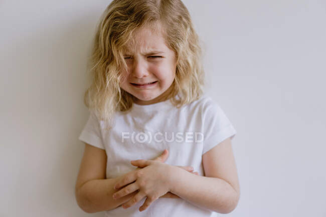 Непослушный ребенок с волнистыми волосами в повседневной одежде стоит со сложенными руками и плачет, глядя в камеру на белом фоне — стоковое фото