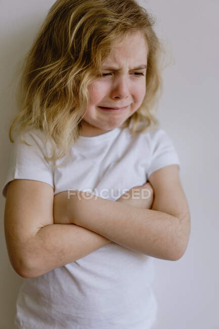 Niño travieso con el pelo ondulado en ropa casual de pie con los brazos cruzados y llorando con los ojos cerrados sobre fondo blanco - foto de stock