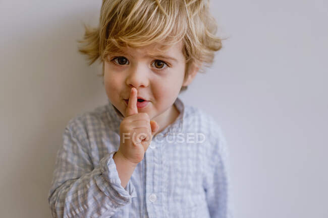 Симпатичный малыш в повседневной одежде стоит на белом фоне студии и прикладывает указательный палец к губам, глядя в камеру — стоковое фото