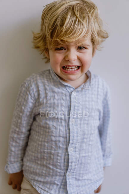 Deluso bambino in camicia casual in piedi vicino al muro bianco e piangendo su sfondo bianco in studio — Foto stock