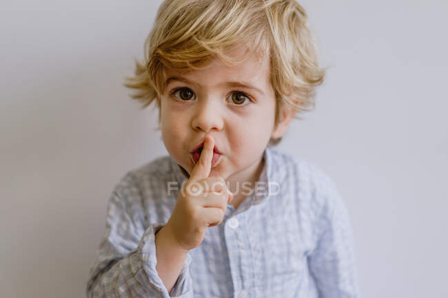 Lindo niño pequeño con ropa casual de pie sobre fondo blanco del estudio y poner el dedo índice en los labios mientras se mira a la cámara - foto de stock