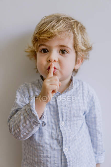 Nettes kleines Kind in lässiger Kleidung steht auf weißem Hintergrund des Studios und legt den Zeigefinger auf die Lippen, während er in die Kamera schaut — Stockfoto