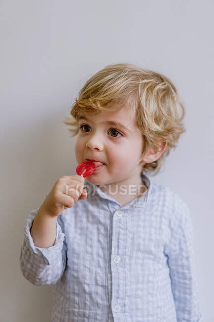 Criança adorável com cabelo loiro desfrutando saboroso pirulito vermelho enquanto está de pé contra a parede cinza — Fotografia de Stock