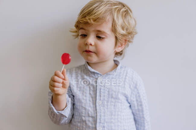Adorable toddler with blond hair enjoying tasty red lollipop while standing against gray wall — Stock Photo