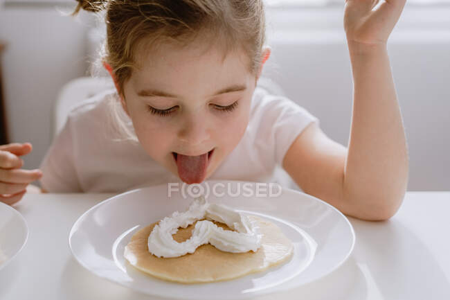 Возбужденный маленький ребенок в повседневной футболке сидит за столом с тарелкой вкусного блина, украшенного взбитыми сливками в форме сердца — стоковое фото