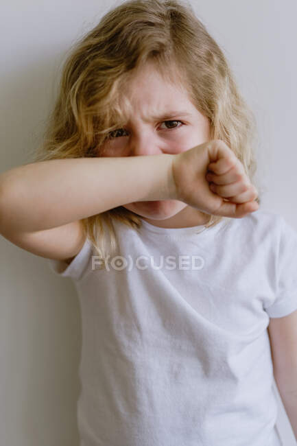 Niño travieso con el pelo ondulado en ropa casual de pie con los brazos cruzados y llorando mirando a la cámara en el fondo blanco - foto de stock