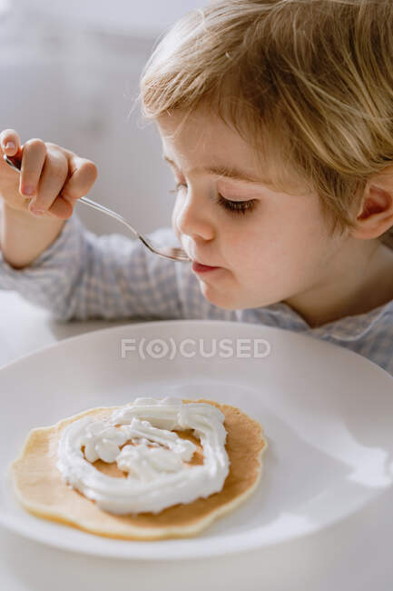 Criança adorável comendo deliciosa panqueca decorada com chantilly enquanto sentado à mesa na cozinha brilhante e tomando café da manhã — Fotografia de Stock