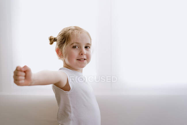 Вид збоку вмісту маленька дитина в повсякденному вбранні стоїть у світлій квартирі і розтягує руки під час прогріву, дивлячись на камеру — стокове фото