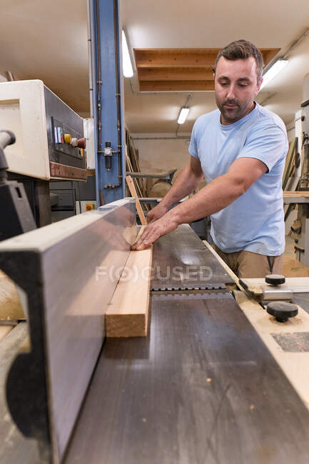 Travailleur du bois dans des vêtements décontractés mettant au point et coupant le bois à l'aide d'une machine électrique spéciale tout en travaillant dans un atelier moderne léger — Photo de stock
