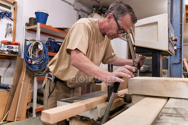 Baixo ângulo de woodworker masculino atencioso de meia idade em óculos e roupas casuais com foco e perfuração de madeira serrada usando máquina elétrica especial enquanto trabalhava na oficina contemporânea — Fotografia de Stock