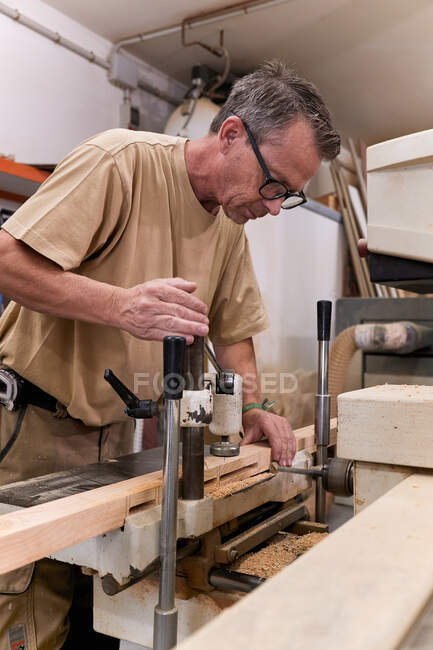 Niedriger Winkel des aufmerksamen männlichen Holzarbeiters mittleren Alters in Brille und lässiger Kleidung, der mit einer speziellen elektrischen Maschine Holz konzentriert und bohrt, während er in einer modernen Werkstatt arbeitet — Stockfoto