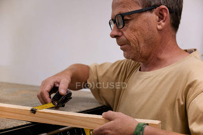 Trabajador de carpintería enfocado en los cultivos en gafas y ropa casual controlando el tamaño del detalle de madera usando cinta métrica mientras trabaja en taller moderno de luz - foto de stock