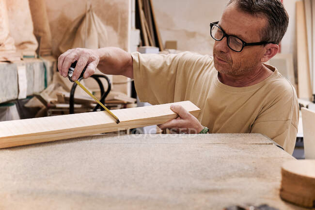 Работник столярного дела в очках и повседневной одежде, управляющий размером деревянных деталей с помощью измерительной ленты во время работы в современной светлой мастерской — стоковое фото