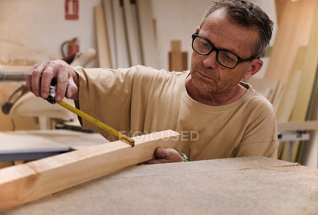 Lavoratore di falegnameria in occhiali e abiti casual che controlla la dimensione del dettaglio in legno utilizzando il nastro di misurazione mentre lavora in officina leggera moderna — Foto stock
