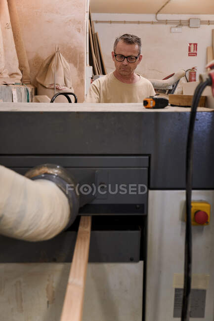 Працівник середнього віку в окулярах і повсякденному одязі, фокусуючись і використовуючи електричну машину, працюючи з деревиною в сучасній теслярській студії. — стокове фото
