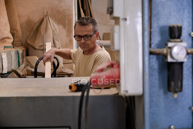 Attentive middle aged workman in glasses and casual clothes focusing and using electric machine while working with timber in light modern carpentry studio — Stock Photo