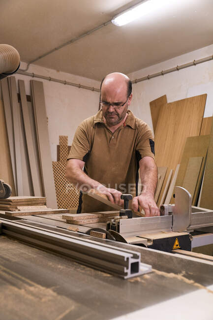 Bajo ángulo de carpintero masculino enfocado en gafas y ropa casual aserrando madera usando una máquina de recorte eléctrica especial mientras trabaja en un estudio de carpintería moderno y ligero - foto de stock