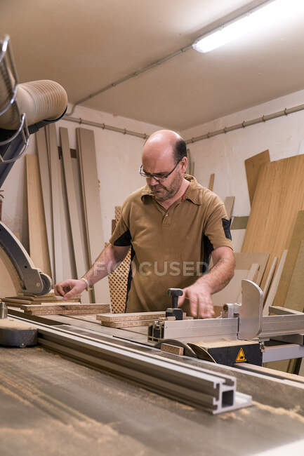 Low angle of focused male woodworker in glasses and casual clothes sawing lumber using special electric trimming machine while working in light modern carpentry studio — Stock Photo