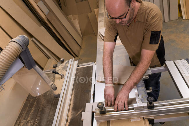 From above crop serious focused carpentry master in glasses and casual clothes measuring and sawing timber while crafting detail using electric trimming machine in light contemporary workshop — Stock Photo