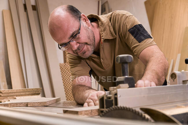 Maestro de carpintería en gafas y ropa casual midiendo y aserrando madera mientras elabora detalles utilizando una máquina de recorte eléctrica en un taller contemporáneo ligero - foto de stock