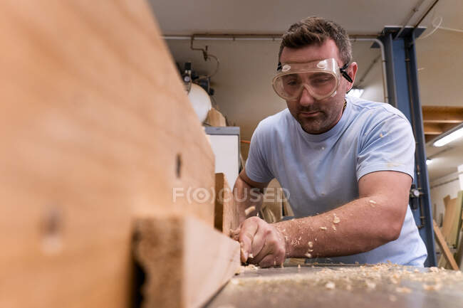 Bajo ángulo de carpintero enfocado en gafas protectoras fresando madera mientras se elaboran detalles utilizando la máquina eléctrica en taller moderno ligero - foto de stock