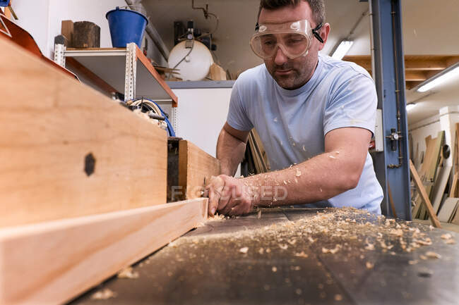 Baixo ângulo de carpinteiro focado em óculos de proteção moagem de madeira enquanto elabora detalhes usando máquina elétrica na oficina moderna leve — Fotografia de Stock