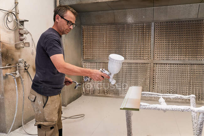 Side view of serious middle aged carpenter using spray gun for painting wood detail with white foamy flame retardant while working in light modern workroom — Stock Photo