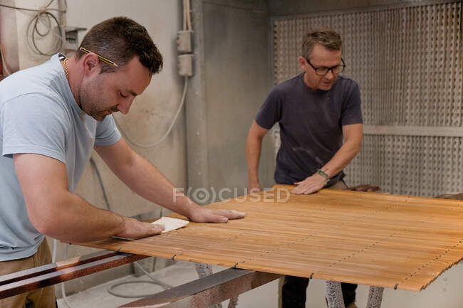 Erwachsener fokussierter Mann poliert mit Lack bemalte Holzlatten, während er mit Kollegen am Regal in der modernen Tischlerei arbeitet — Stockfoto