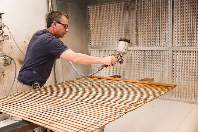 Adulto focado homem usando pistola de pulverização pintura ripas de madeira com laca enquanto trabalhava na sala de trabalho de carpintaria contemporânea — Fotografia de Stock