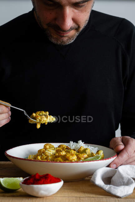 Male in casual wear sitting at table in kitchen and taking piece of spicy curry made of prawns and mushrooms and served with rice and lime slices — Stock Photo