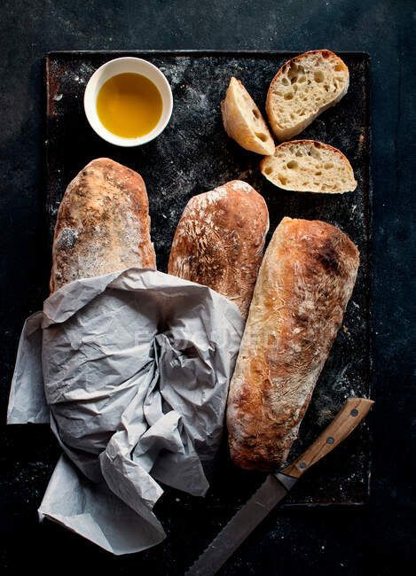 Pão Ciabatta em tábua rústica perto de azeite e faca com fatias de pão no fundo escuro — Fotografia de Stock