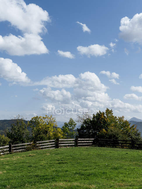 Increíble paisaje de zona montañosa con exuberante prado rodeado de barrera de madera bajo el cielo azul en verano - foto de stock