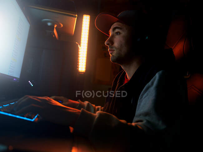 Вид сбоку сфокусированного мужчины в кепке и беспроводной гарнитуре в случайной одежде, сидящего в одиночестве за компьютером и играющего в видеоигру в темной комнате с тусклым синим светом ночью — стоковое фото
