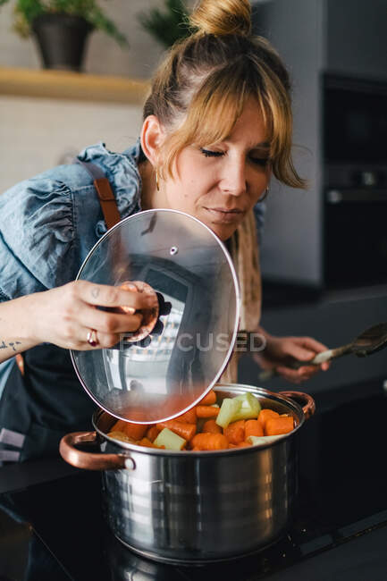 Tranquilo fêmea em pé avental com os olhos fechados perto fogão na cozinha contemporânea e desfrutar de sabor de legumes aromáticos preparados em panela — Fotografia de Stock