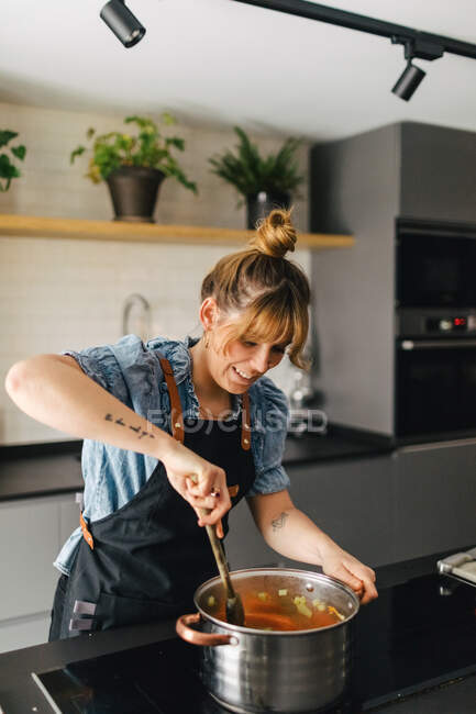 Стильна жінка в фартусі стоїть біля плити і перемішує інгредієнти в каструлі під час приготування вечері на сучасній кухні — стокове фото