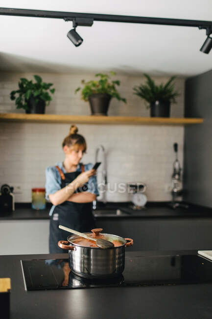 Elegante dona de casa vestindo avental moderno inclinado no balcão na cozinha e navegando telefone celular enquanto espera pela preparação de delicioso jantar na panela no fogão — Fotografia de Stock