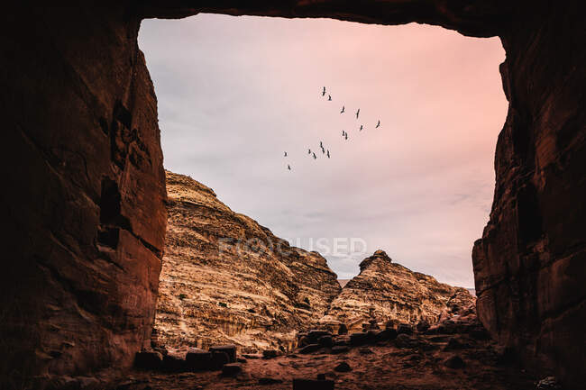 Amazing view of rocky mountains and flock of flying birds on pink cloudy sky during sunset from inside cave entrance — Stock Photo