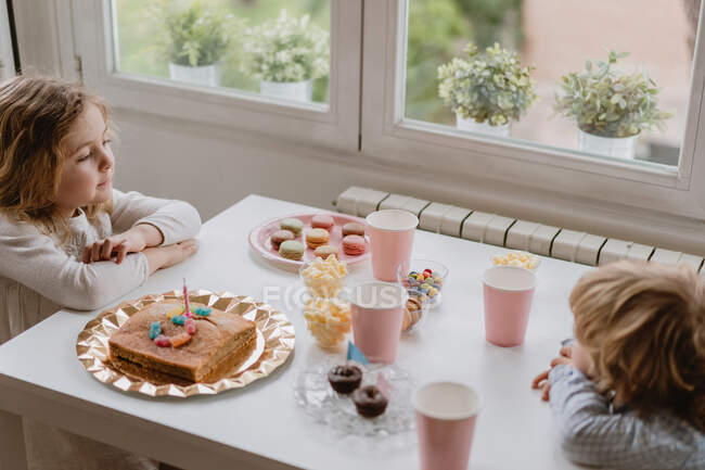 From above of cheerful little kids in casual clothes sitting at wooden table near window and eating sweet cake during holiday — Stock Photo