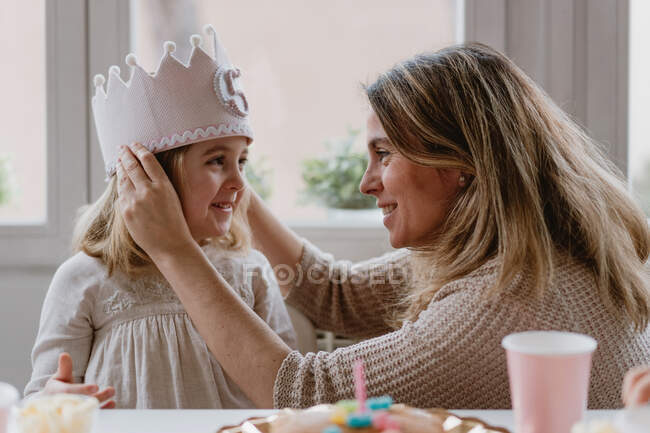 Vista lateral de la alegre madre colocando fieltro corona hecha a mano en la hija mientras celebran cumpleaños juntos en casa - foto de stock