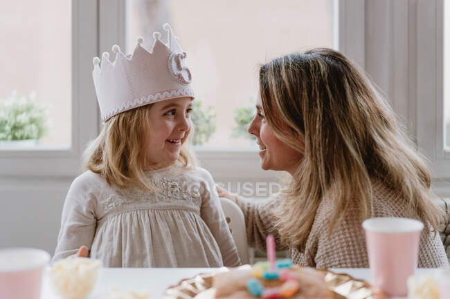 Vue latérale de la mère gaie plaçant la couronne faite main de feutre sur la fille tout en célébrant l'anniversaire ensemble à la maison — Photo de stock