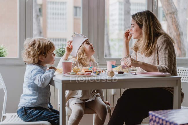 Positive mother in casual clothes sitting at wooden table and talking with kids celebrating birthday at home — Stock Photo