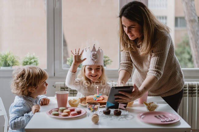 Positive mother and daughter in casual clothes sitting together at table and making video call on tablet while celebrating birthday at home — Stock Photo