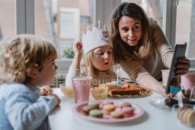 Madre e hija positivas en ropa casual sentadas juntas en la mesa y haciendo videollamada en la tableta mientras celebran su cumpleaños en casa - foto de stock