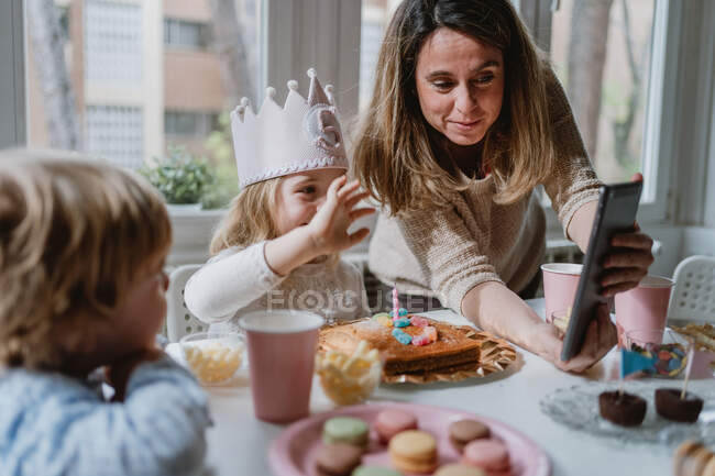 Madre e hija positivas en ropa casual sentadas juntas en la mesa y haciendo videollamada en la tableta mientras celebran su cumpleaños en casa - foto de stock