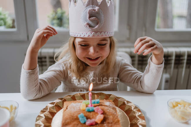 Bonne petite fille en vêtements décontractés et avec couronne en feutre soufflant bougie sur le gâteau d'anniversaire tout en étant assis à la table en bois pendant la fête — Photo de stock